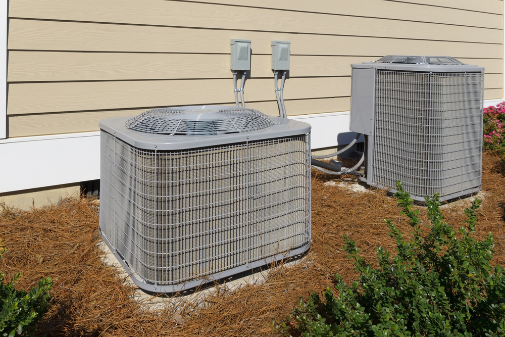 Two outdoor air conditioning units installed on the ground beside a beige building with electrical boxes mounted on the wall.