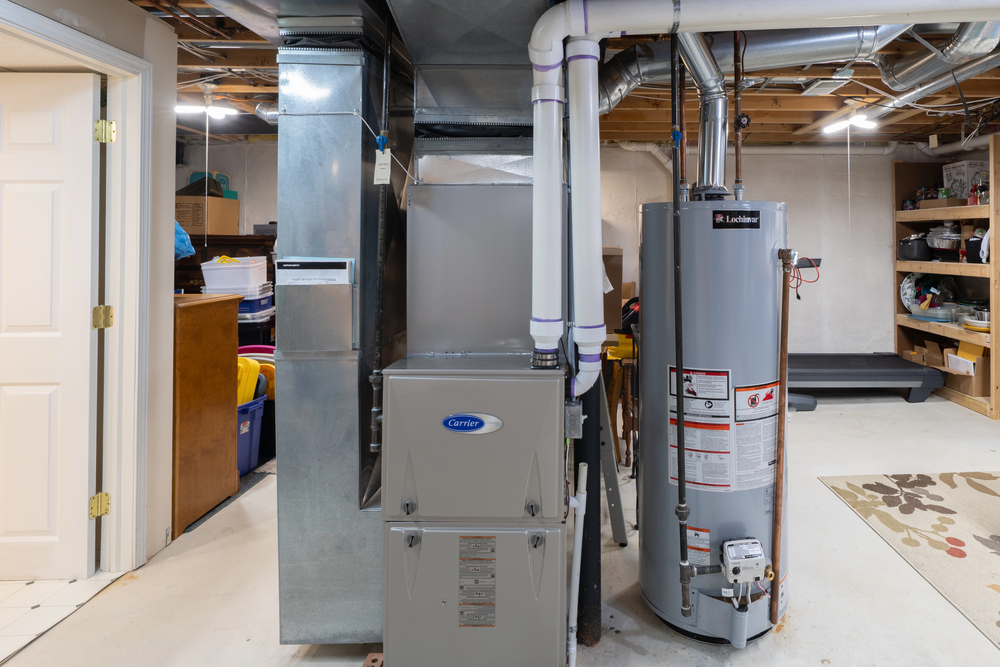 A basement utility area with a central heating furnace, water heater, exposed pipes, shelves with storage items, and a treadmill in the background.