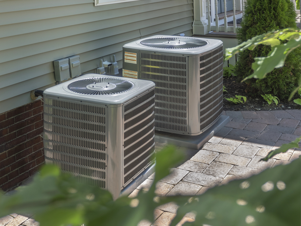 Two outdoor air conditioning units are placed on a paved area beside a house with vinyl siding and brick foundation; some greenery is visible in the foreground.