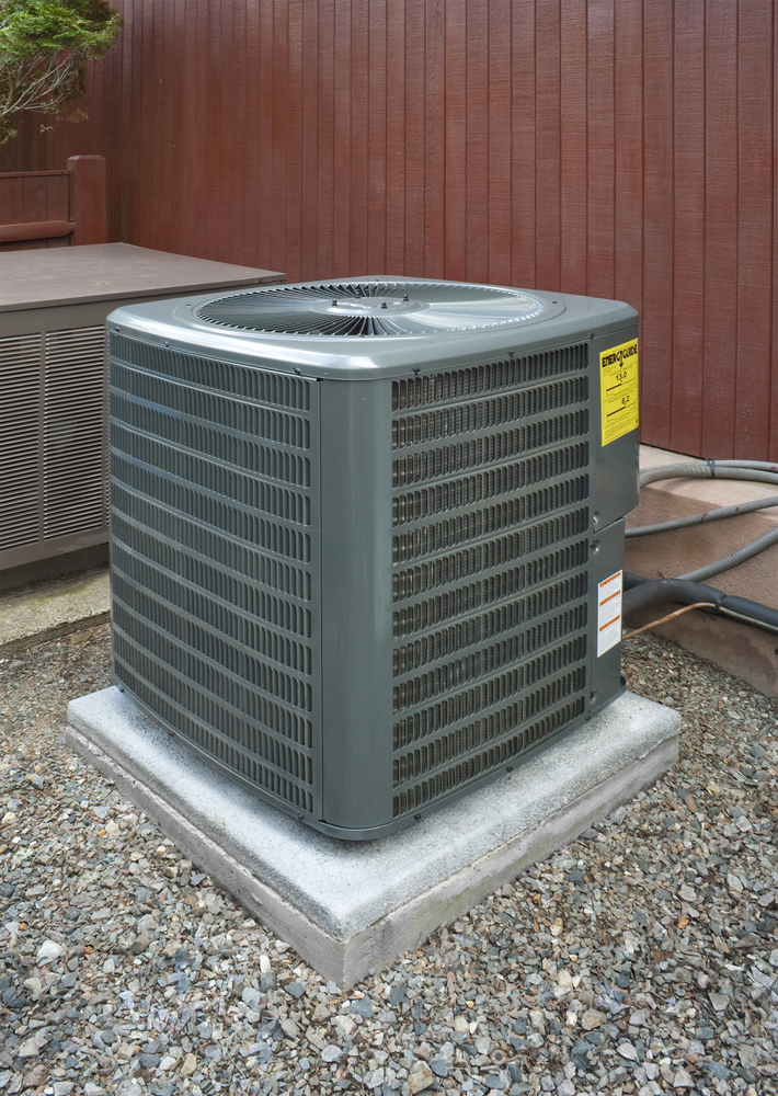 A central air conditioning unit sits on a concrete pad surrounded by gravel, positioned next to a wooden fence and connected to nearby piping.