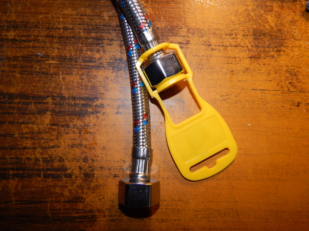 A braided metal hose with a hexagonal connector next to a yellow plastic tool on a wooden surface.