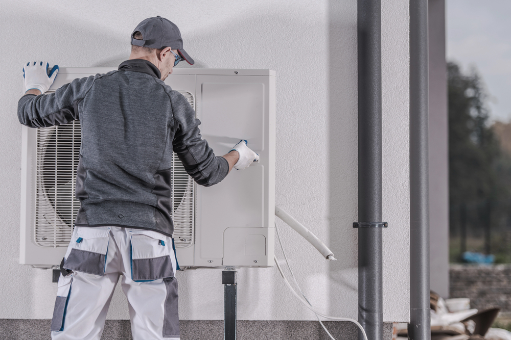A technician in work attire inspects and services an outdoor air conditioning unit mounted on a building wall.