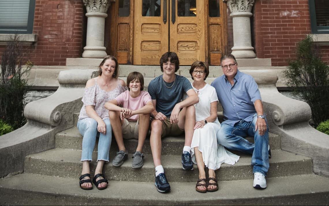Five people sit together on stone steps in front of a wooden double door, posing for a group photo in casual attire.
